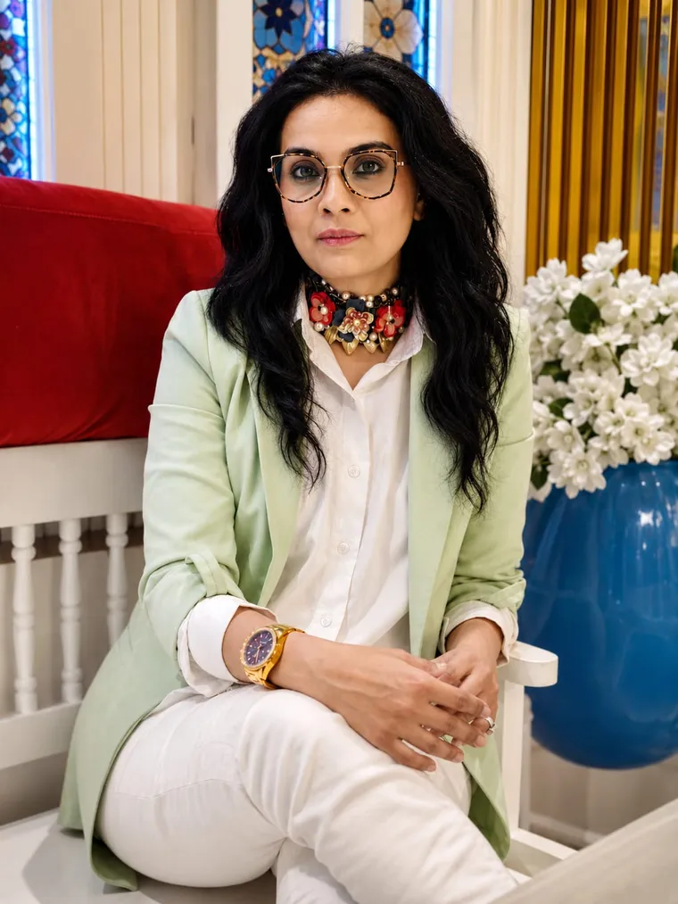 Maliha Bano seated in a sage blazer beside a blue vase of white flowers